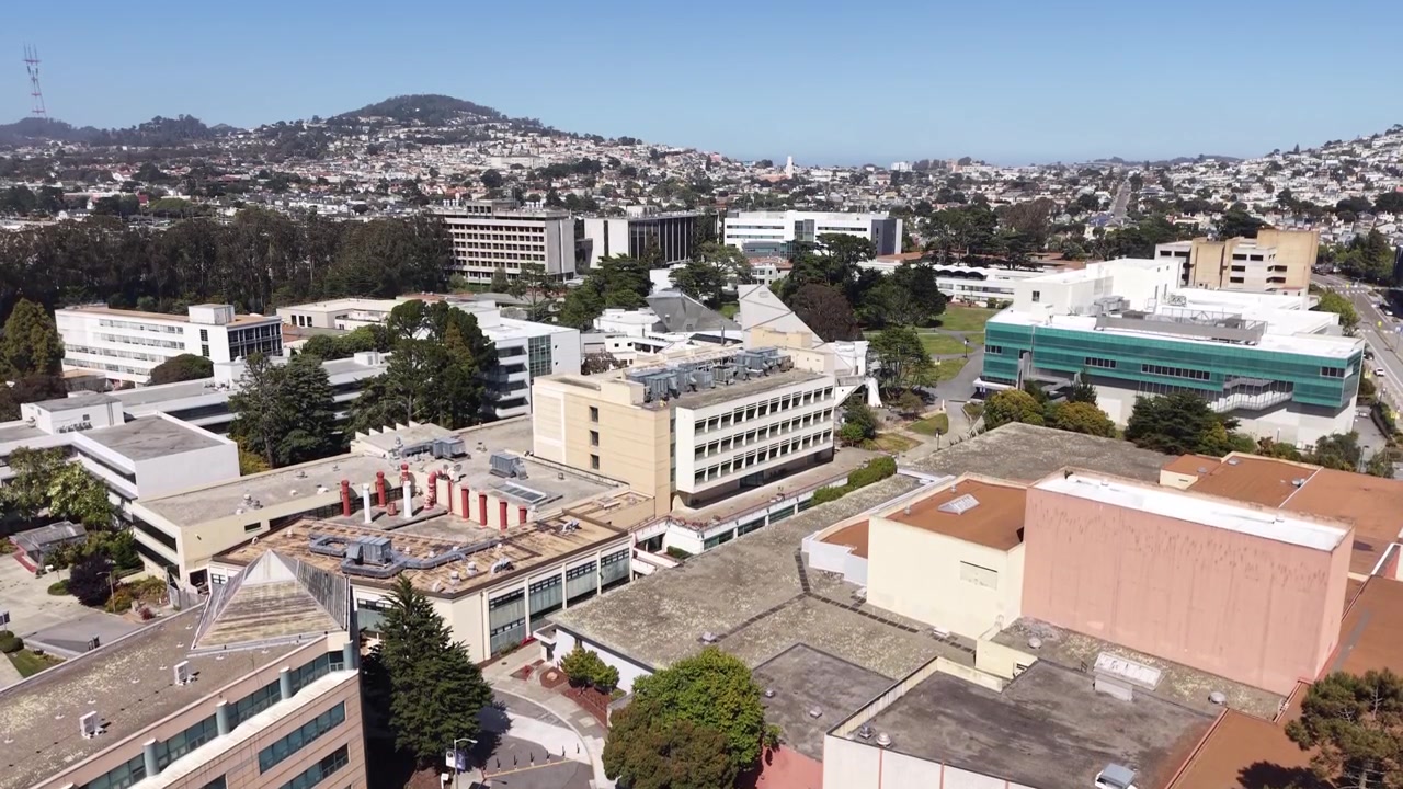 Rekaman drone sinematik yang terbang perlahan di atas bangunan modern dan ruang hijau kampus Universitas Negeri San Francisco pada hari yang cerah dan terang, dengan perbukitan kota di latar belakang.