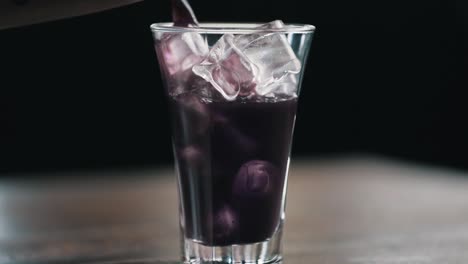 a purple drink is poured into a glass filled with ice cubes, close-up view from the side