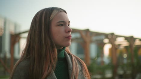 lady in gray coat holding book studies reflectively outdoors, gazing thoughtfully into distance with calm expression, wooden pergola and modern residential buildings blurred in background