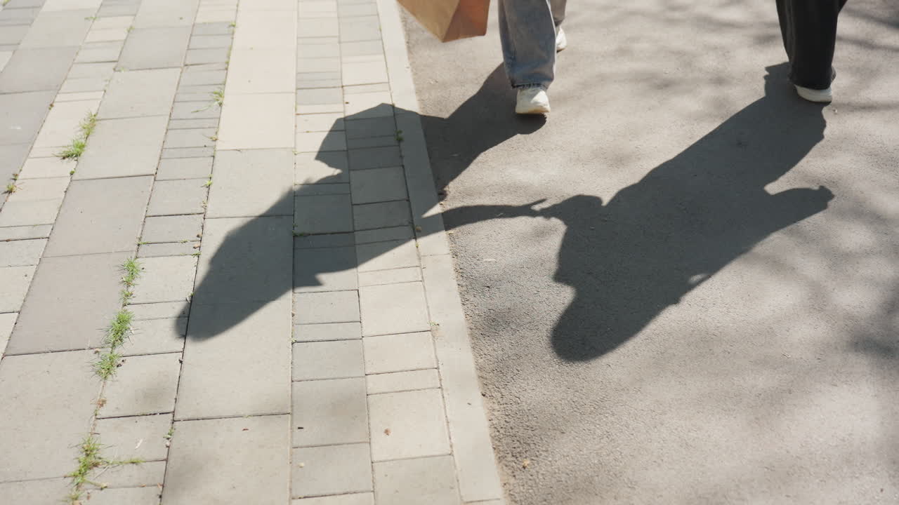 타일이 깔린 보도를 걷는 보행자들이 손을 잡고 있는 모습, 선명한 햇빛이 긴 실루엣을 드리우고 질감이 느껴지는 포장 도로, 쇼핑백을 들고 활기차게 걷는 모습과 도시의 리듬, 찰나의 순간