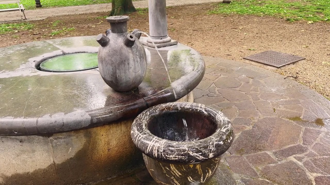 Närbild av en skulptural offentlig dricksvattenfontän i en park i Ljubljana, med vatten som rinner från ett urnformat kärl ner i ett väderbitet stenfat