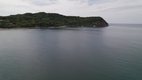 Drone-heads-towards-the-rocky-shores-of-Punta-de-Mita-from-the-sea,-revealing-Careyeros-Beach's-lush-landscape