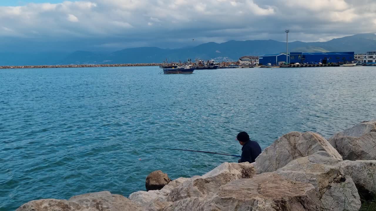 single man fishing at the shore of wavy Mediterranean sea on a cloudy overcast day with harbor of Bejaia and mountains in the background. North Africa Algeria