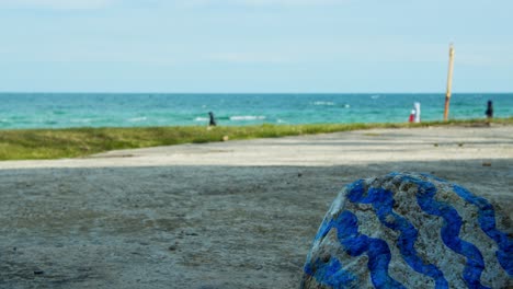 ángulo-Bajo-Cerca-De-Una-Piedra-Pintada,-El-Mar-Negro-En-El-Fondo-Se-Enfoca,-Gente-Caminando-En-La-Playa