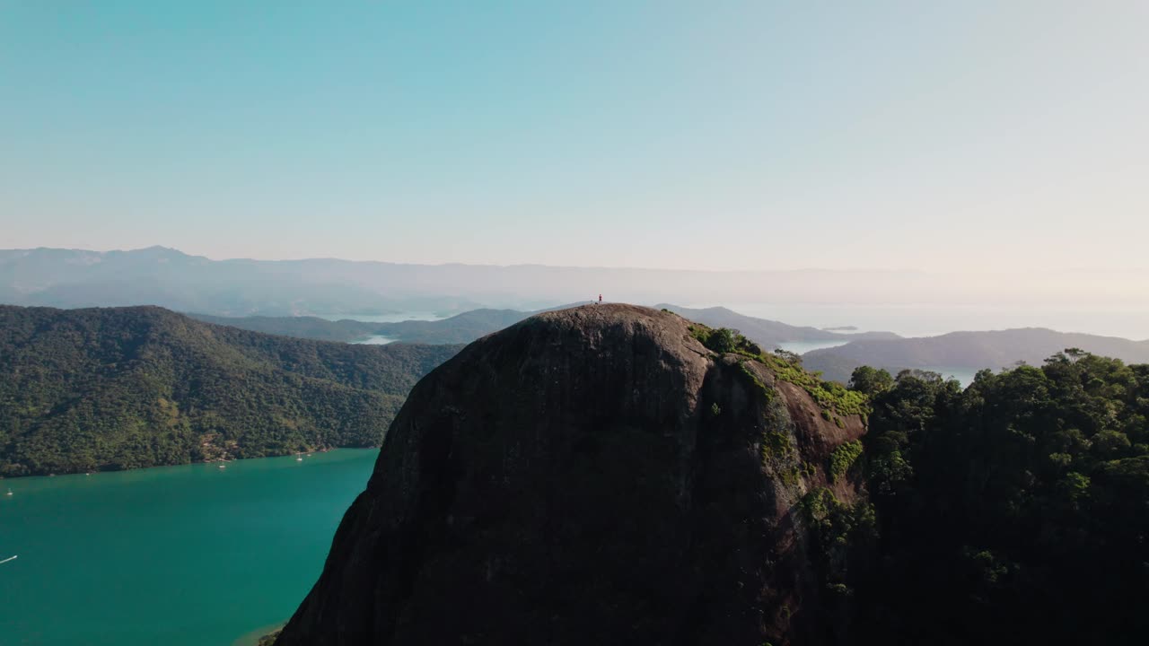 Breathtaking high-angle view of a solitary figure standing atop a huge granite rock formation. Below, clear turquoise water meets lush tropical mountains shrouded in mist