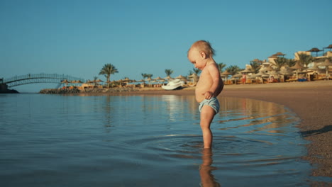 adorable toddler splashing water in warm sea at sand beach at summer holiday.