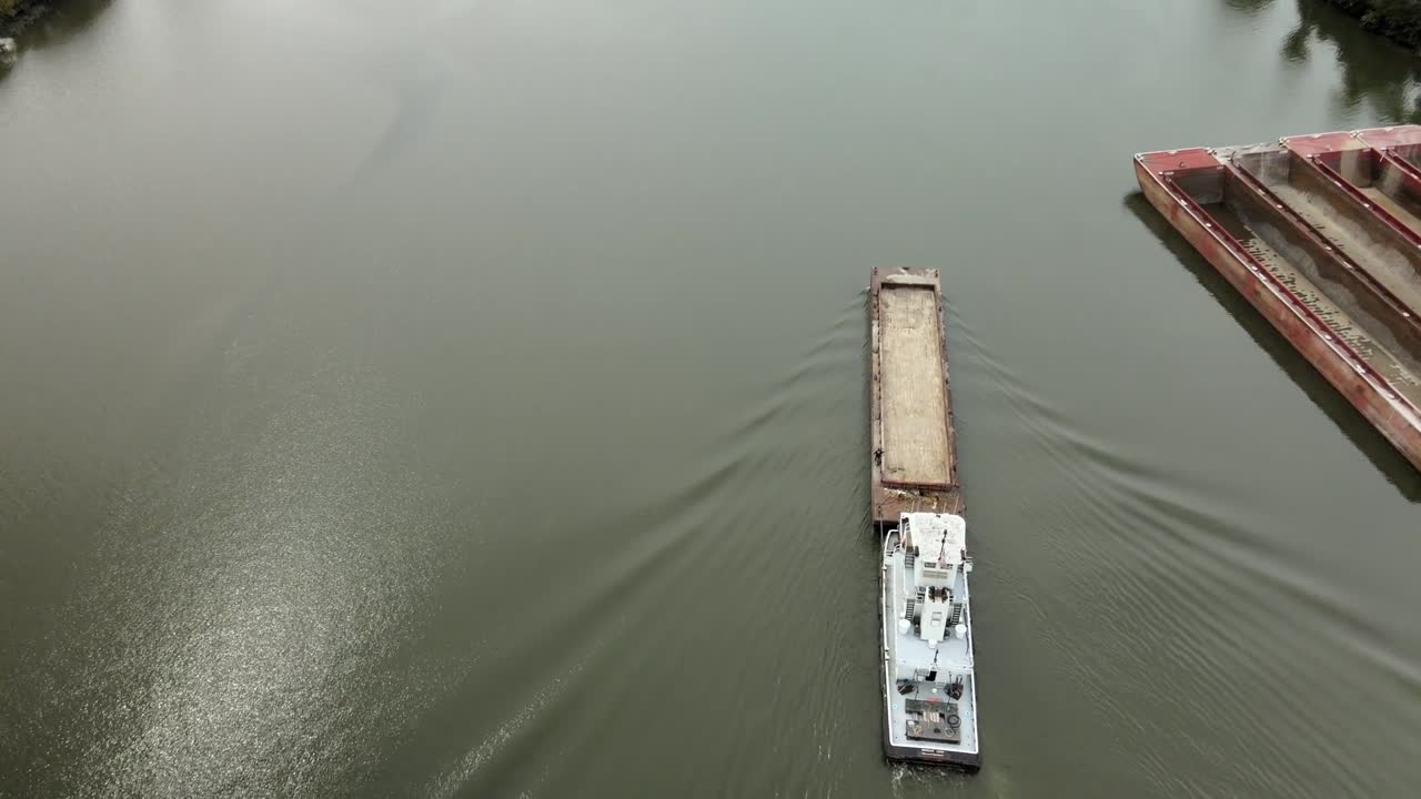 Aerial drone footage of towboat guiding cargo barge along calm Kanawha River surrounded by forested hills, industrial docks, and quiet roads in scenic West Virginia, United States