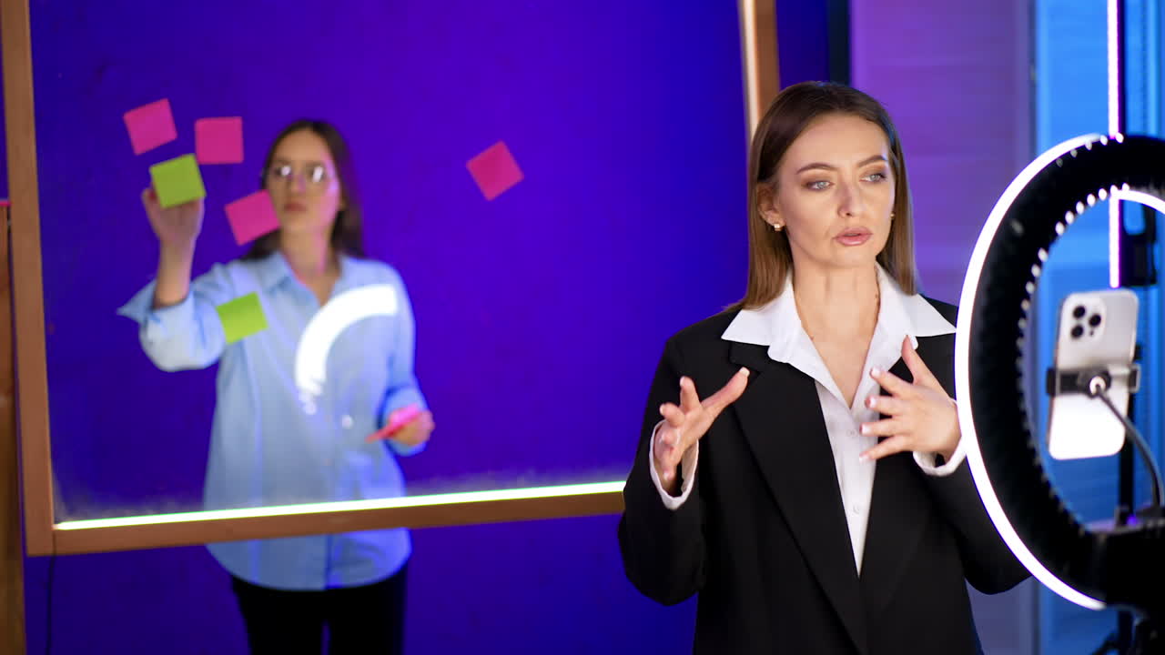 Good-looking Caucasian lady in black jacket takes a video and points at the glass wall behind her. Girl behind the glass board puts stickers on it.