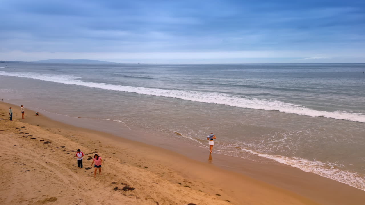 Los Angeles, USA, 29 August 2025: Some people rest at the shore of the Pacific Ocean. Waves roll to the sandy beach on gloomy day. LA, California, USA from drone footage