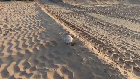 una vieja pelota de fútbol desaliñada rodando a lo largo de la arena de la playa con huellas de neumáticos en la arena en un día soleado