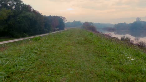 Drone-shot-traveling-along-a-grassy-hill-with-a-nearby-reflective-lake-on-a-foggy-autumn-morning