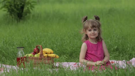 Weekend-at-picnic.-Lovely-caucasian-child-girl-on-green-grass-meadow-sit-on-blanket-waving-her-hands