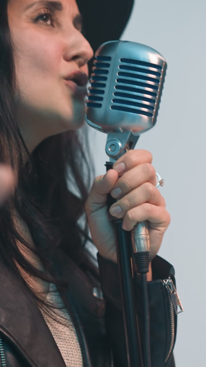 검은 모자와 가죽 재을 입은 열정적인 여성 가수가 빈티지 마이크로 노래하고, 부드러운 색 배경에 대해 표현적으로 움직이고, 카메라 을 통해 근접 뷰
