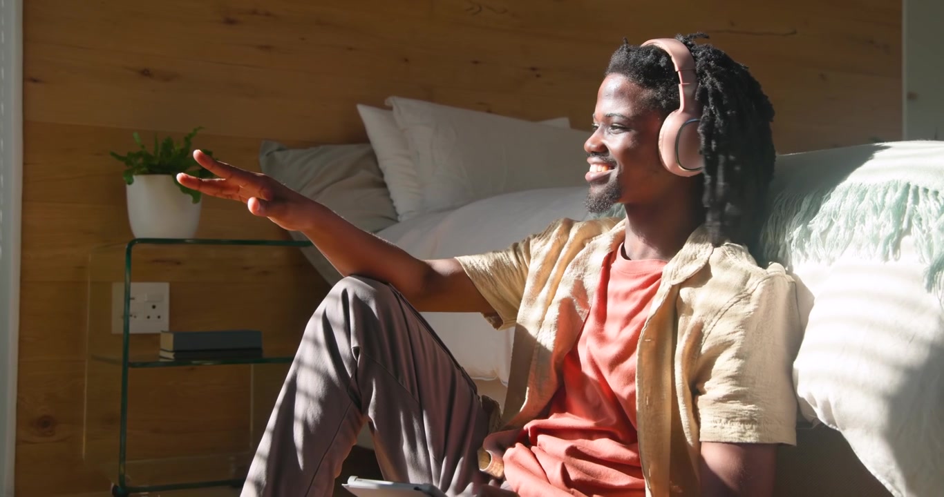Afroameričan sedící v ložnici, držící tablet, s podcastem hrajícím na růžových sluchátkách, usmívající se