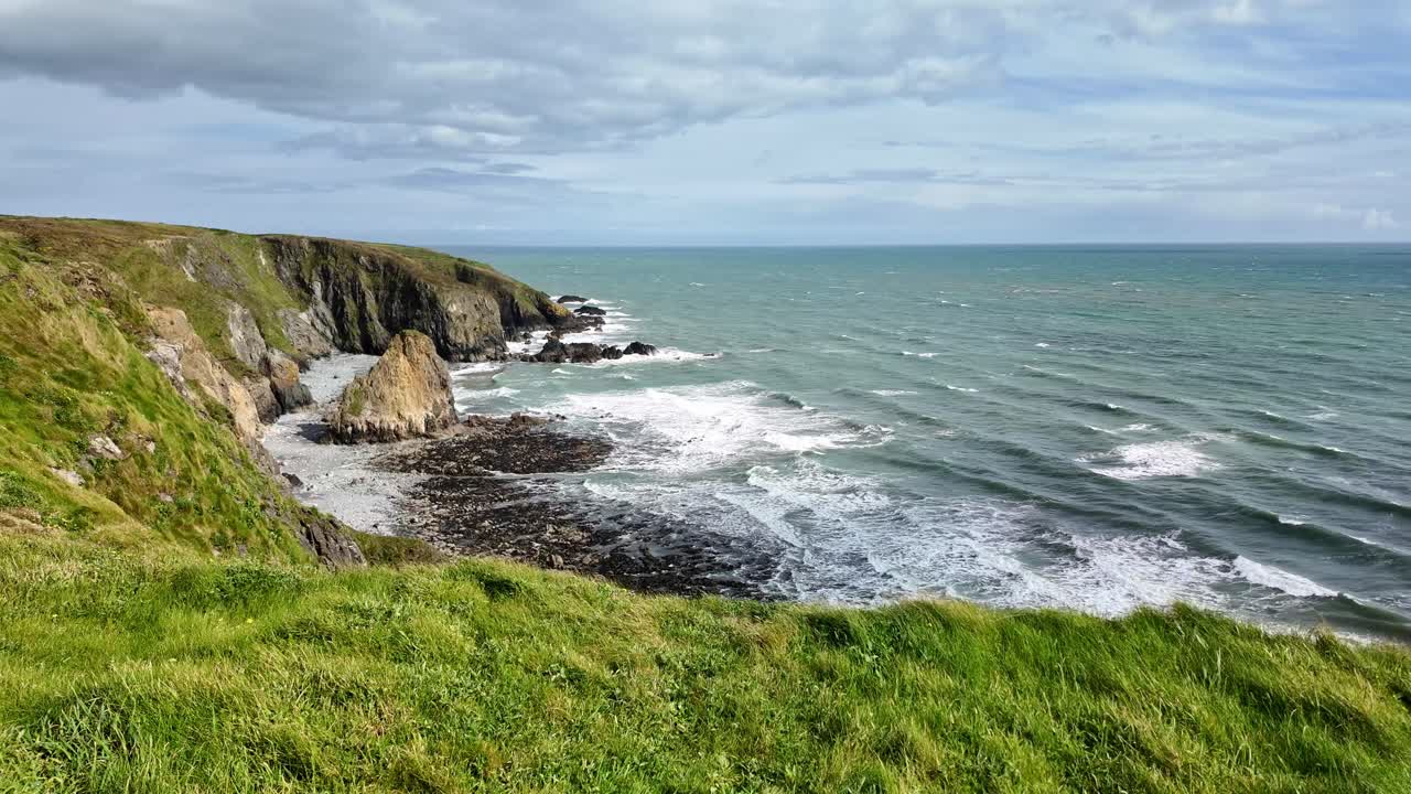 Havlandskab med indkommende tidevand, stor havstabel og frodigt grønt græs på klippekanten ved Copper Coast, Waterford, Irland, en forårseftermiddag i april.