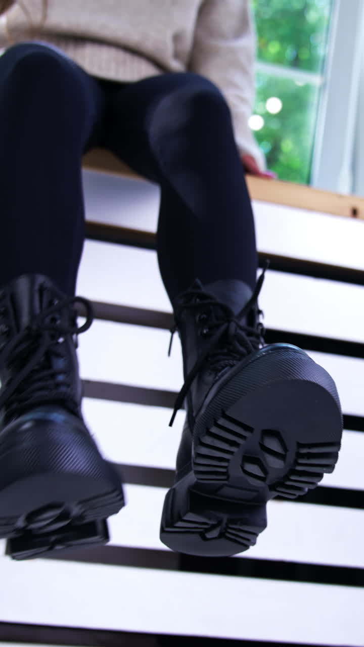 Black boots with laces on tractor soles. Close up. Female model sitting on a window sill demonstrating fashionable footwear. Low angle view. Vertical video