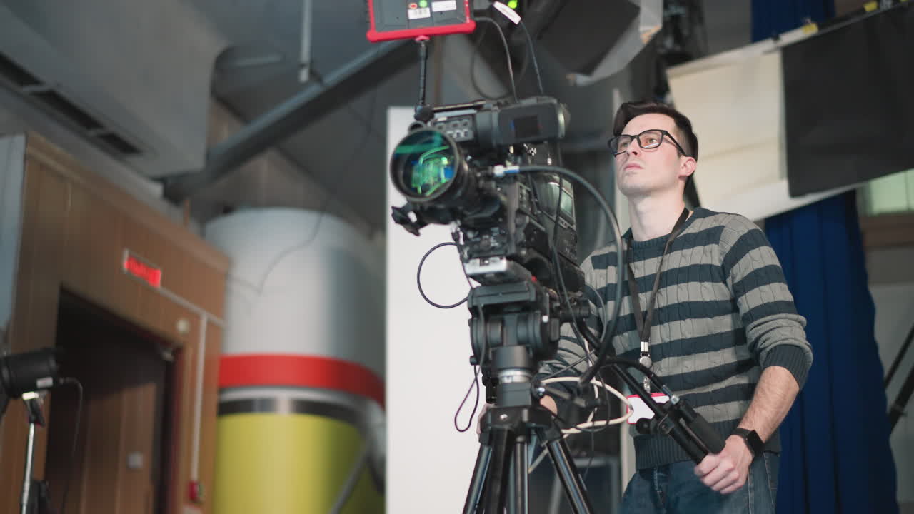 Camera operator adjusting high-end broadcast camera on tripod in studio, focusing on equipment, preparing for shooting scene in professional TV production setup with studio lights