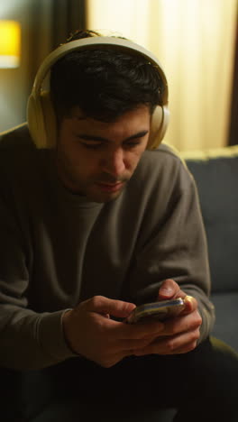 vertical video of man spending evening at home sitting on sofa wearing wireless headphones streaming music or watching content from mobile phone 1