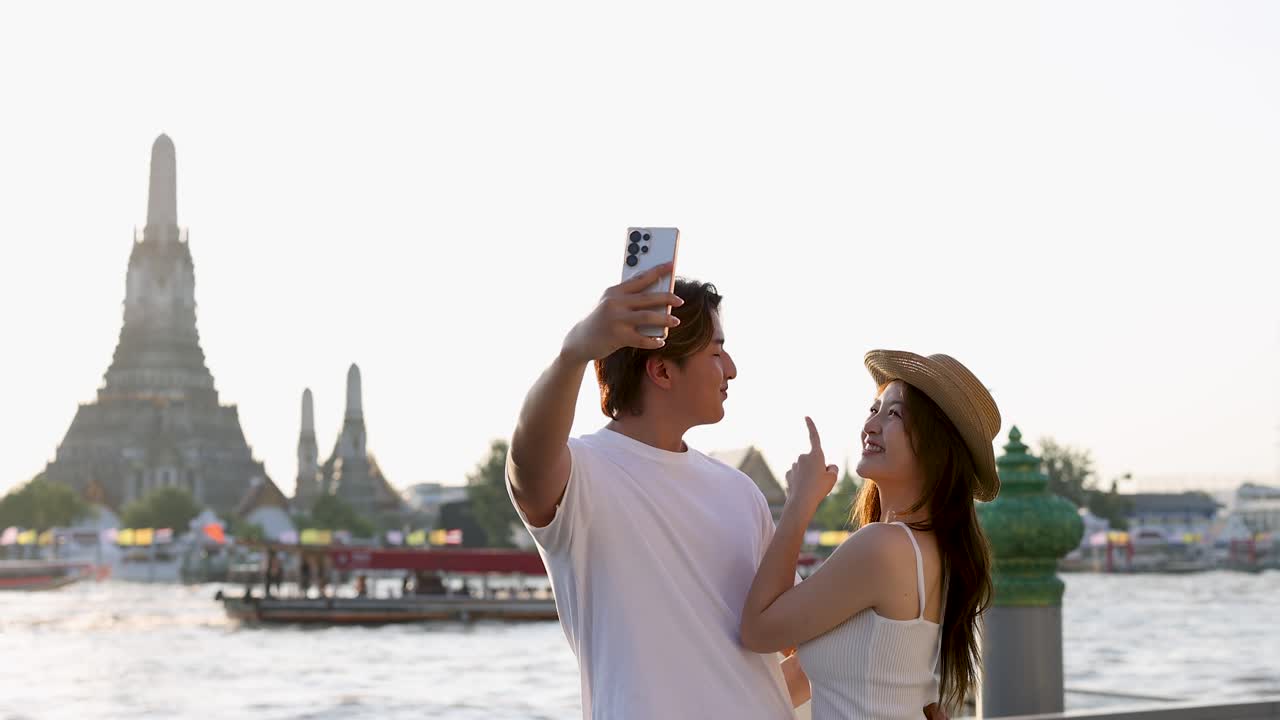 Coppia sorridente che si scatta un selfie vicino a un tempio sul fiume, illuminazione calda del tramonto, atmosfera rilassata da viaggio, camera stabile