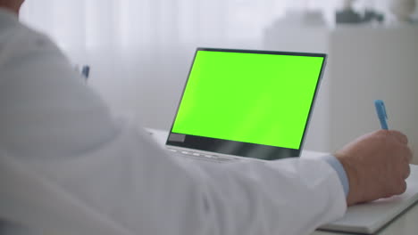 telemedicine-service-consulting-with-best-medical-specialist-online-laptop-with-green-screen-for-chroma-key-technology-on-table-physician-is-writing-notes