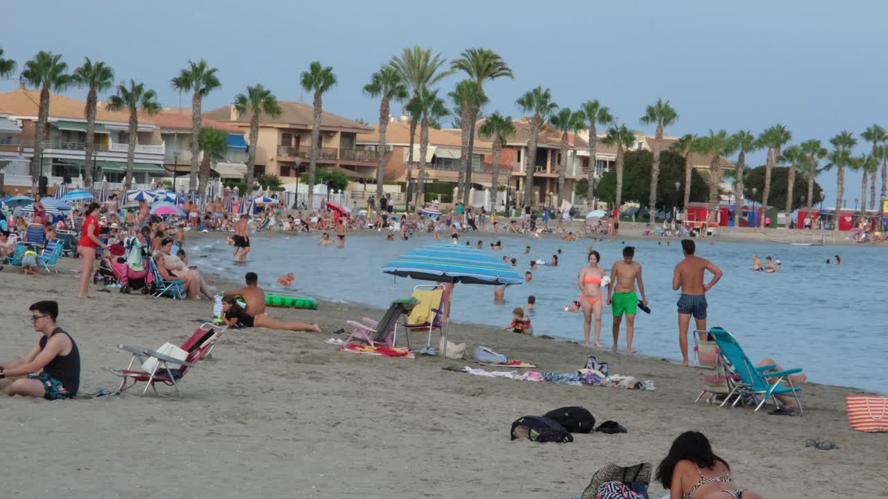 A large crowd enjoys the sunny Los Alcázares beach, relaxing on white sand, swimming, and playing in the warm sea during the vibrant summer months.