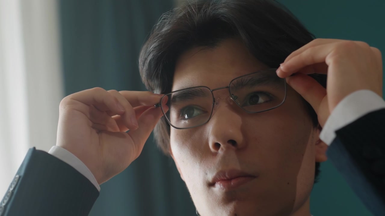 スーツを着た若い男性が眼鏡を置く。落ち着いた表情、最後の身だしなみ、揺るぎない視線で自信を示している。柔らかなカーテンのあるオフィス内、シネマティックな照明、ローアングル、堂々とした佇まい。