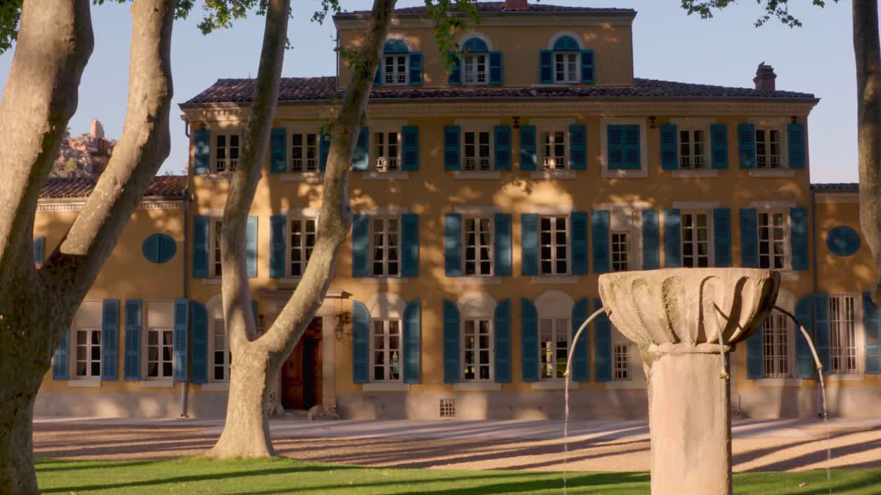 A wide shot pans left across a yellow mansion with blue shutters, surrounded by trees and a stone fountain in a serene garden setting.