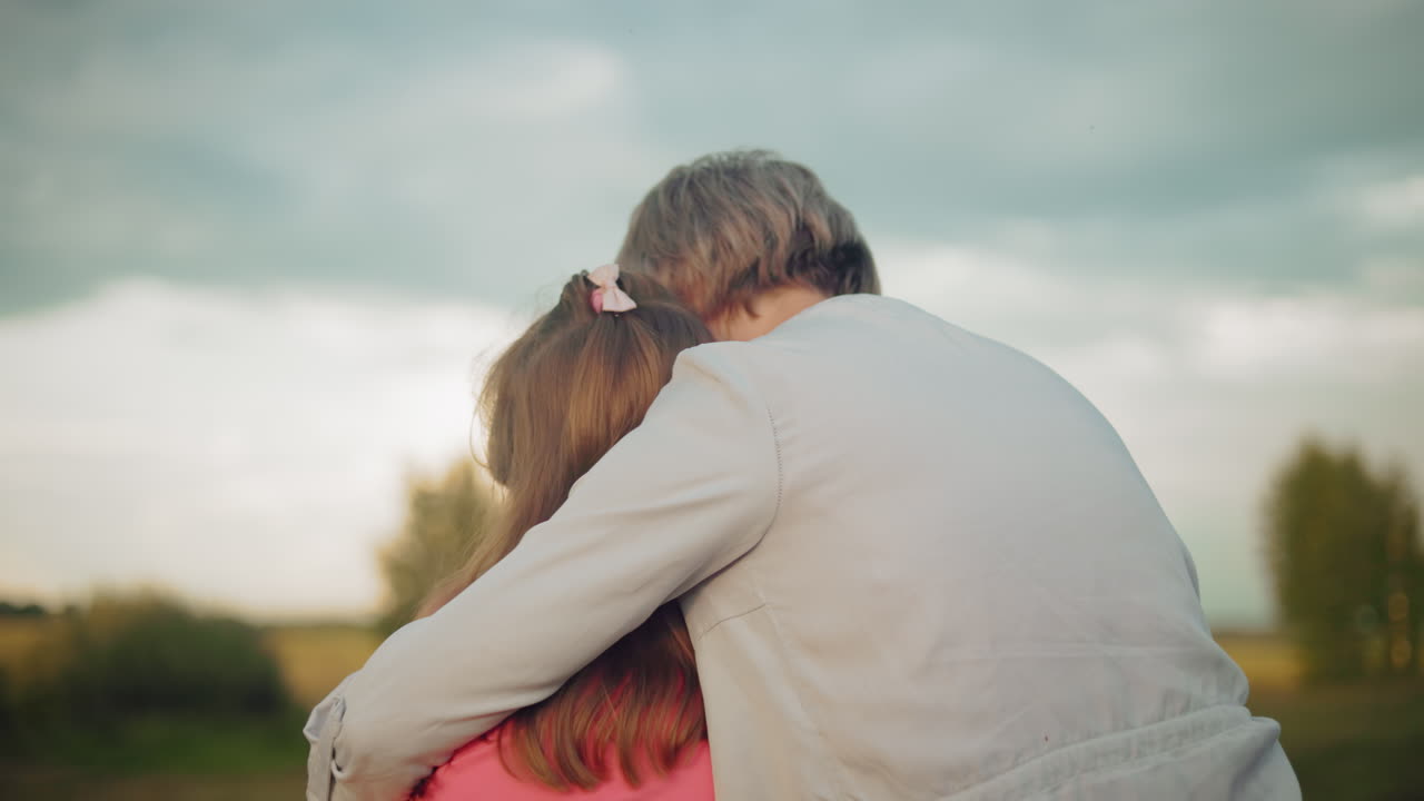momento conmovedor como una madre abraza a su hija en rosa, ambos sentados en una bala de heno, compartiendo la cercanía en un campo expansivo bajo un cielo nublado
