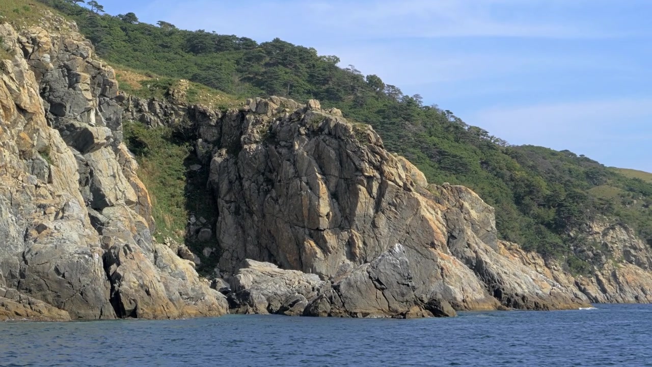 Il passaggio ravvicinato di una barca segue un massiccio promontorio grigio scuro dove un'alta rientranza fratturata forma una grotta marina in ombra sopra rocce costiere frastagliate con bassa vegetazione verde sul pendio superiore