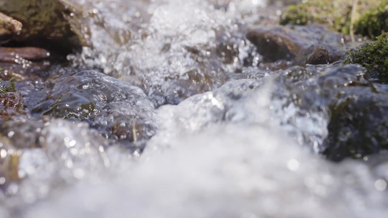 Clear water rushes over mossy stones in a forest stream. Tiny bubbles and splashes shimmer with sunlight. Nature's quiet power moves through rocks and greenery. Ideal for eco themes, wellness