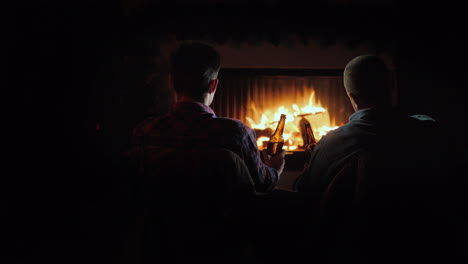 silhouettes of two men by the fireplace relaxing with bottles of beer in their hands