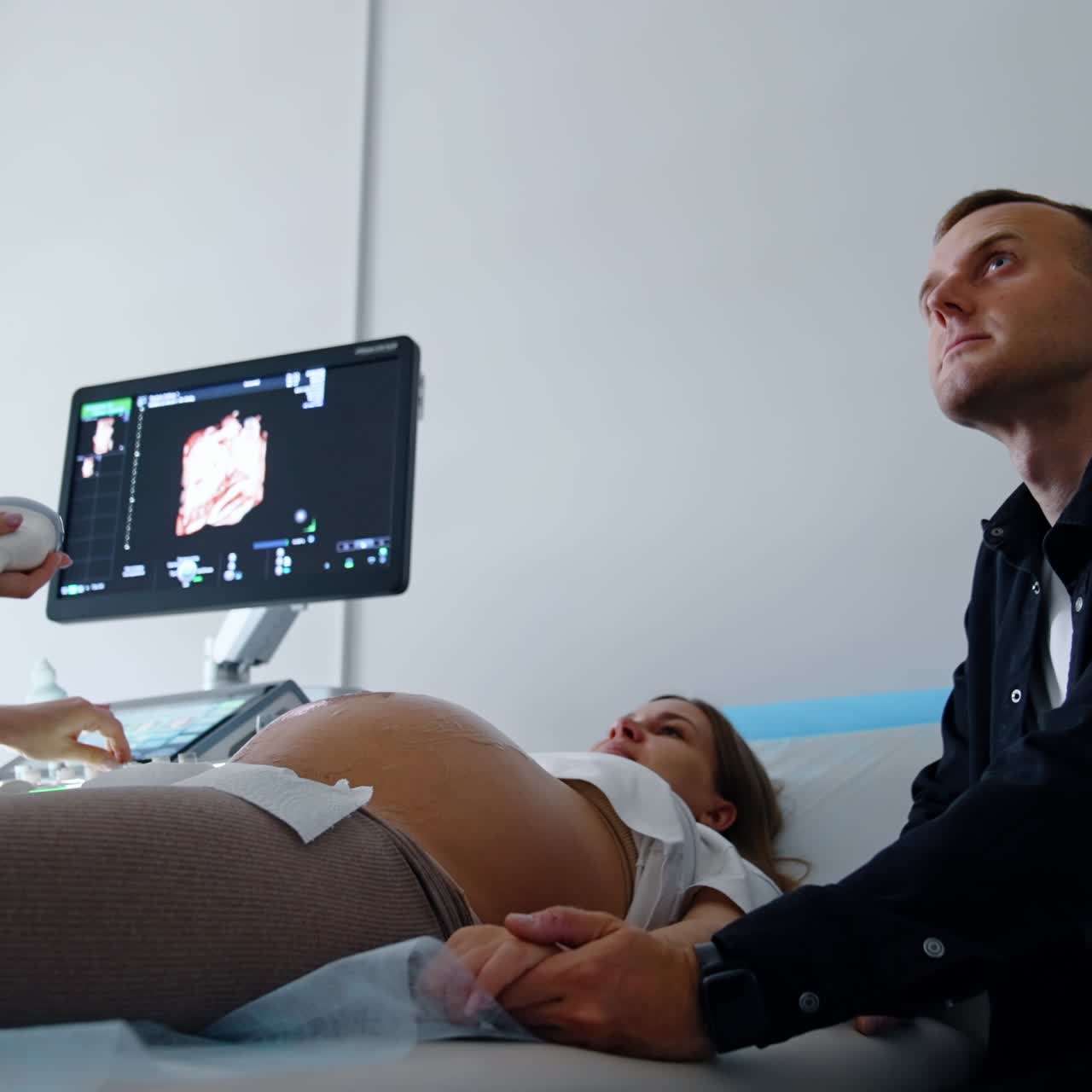 Loving Caucasian couple holding hands in the doctor's cabinet. Pregnant woman is examined at ultrasound machine. Low angle view
