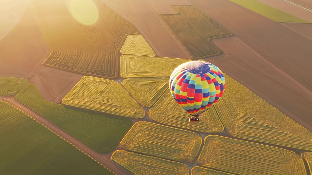 Un pallone luminoso e multicolore si muove costantemente sopra campi agricoli sottostanti illuminati dal sole, mentre lunghi confini dei campi e una leggera foschia dorata creano profondità nella vasta composizione aerea