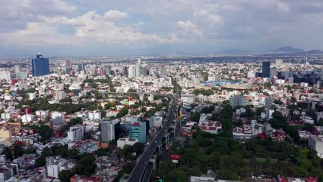 Scenic-flight-above-downtown-Mexico-city-center,-freeway,-cars-traveling-on-expressway,-buildings,-apartments,-towards-Cruz-Azul-stadium-and-mountain-range-in-background,-overhead-aerial-approach