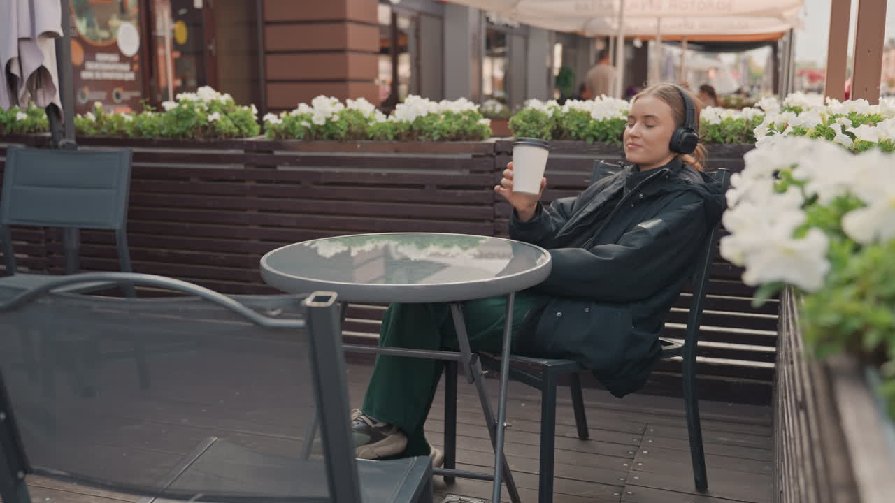 unspecified woman sitting at outdoor cafe sipping coffee from cup while wearing headphones and jacket, leaning back in chair beside planter with white flowers, urban street background, relaxed