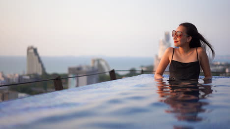 mujer asiática glamorosa sentada en una piscina infinita en la azotea con vista a la ciudad
