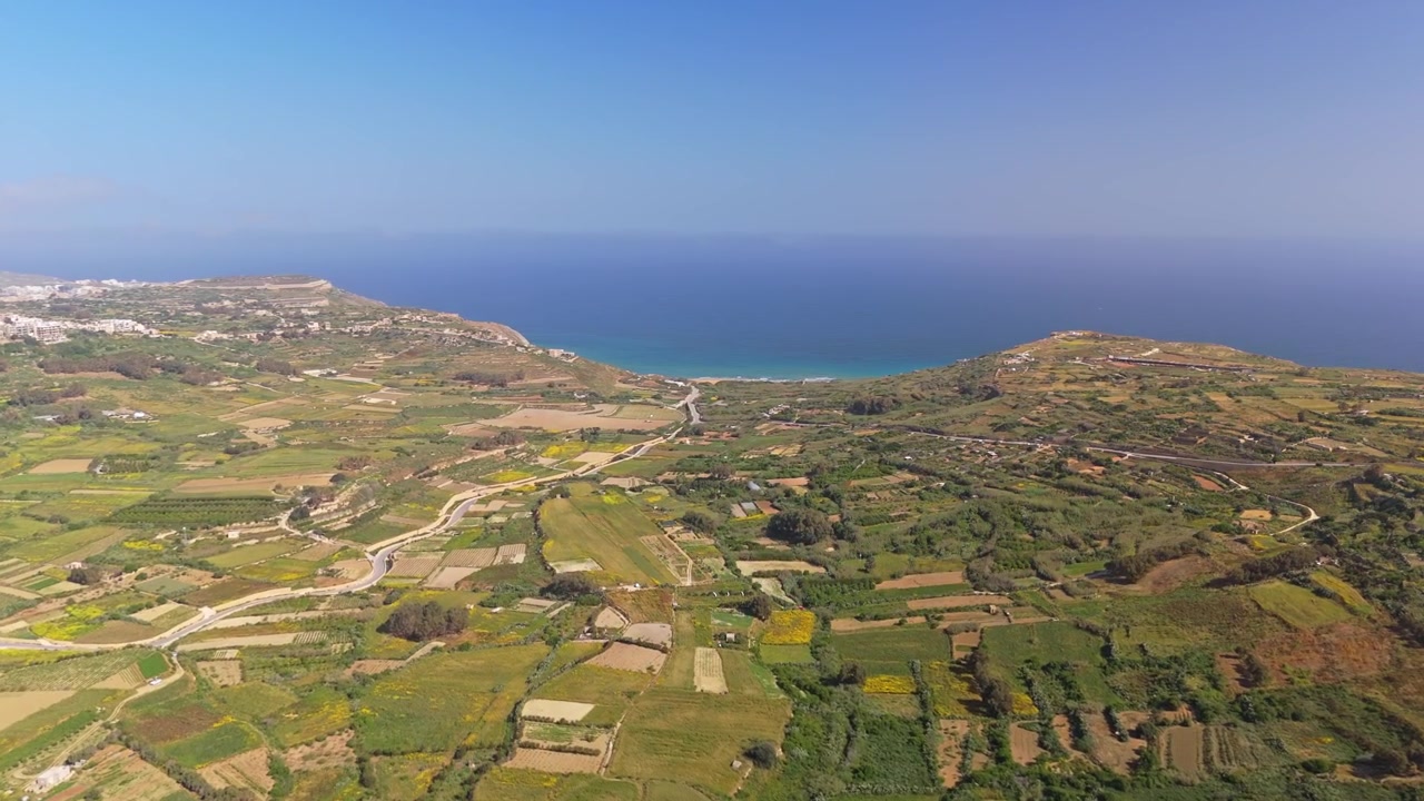 Nella Baia di Ramla a Gozo, una vista aerea mostra la spiaggia e la costa mediterranea delimitate da campi terrazzati e dalla campagna rurale costiera sotto un cielo sereno.