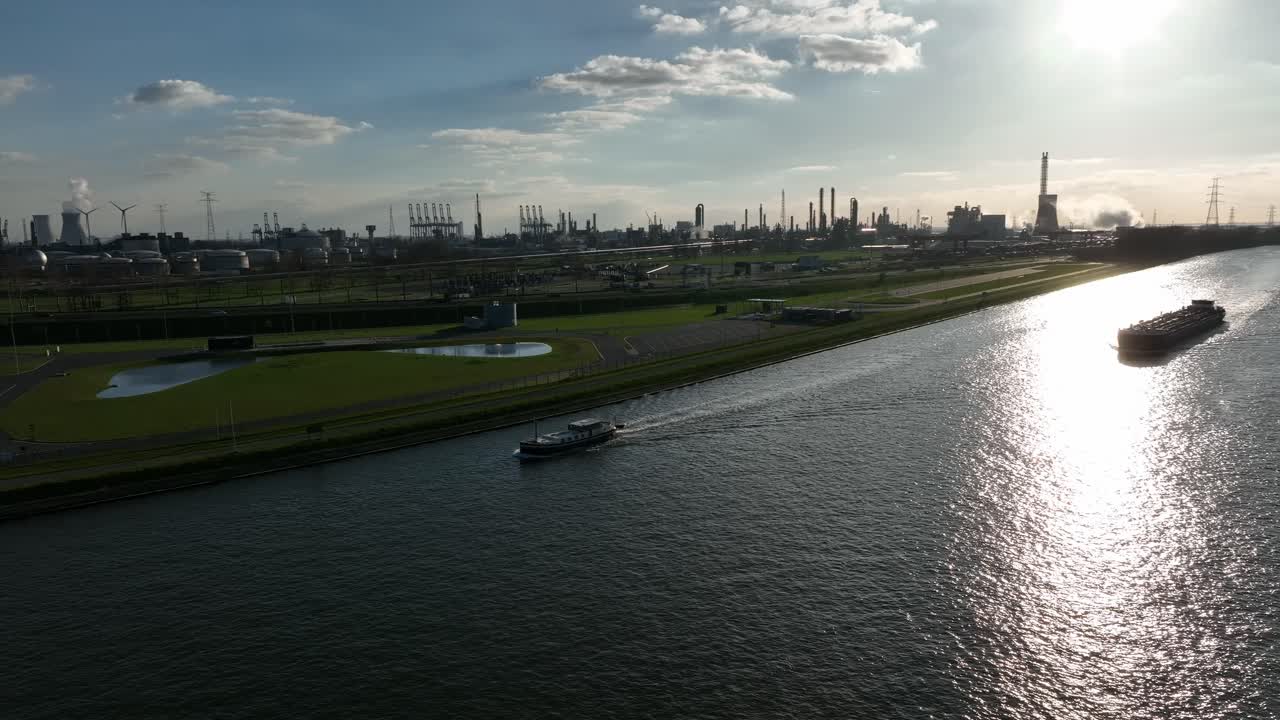 Pan left aerial shot of Antwerp canal with boat navigating alongside factories, pipelines, and wind turbines during late afternoon light