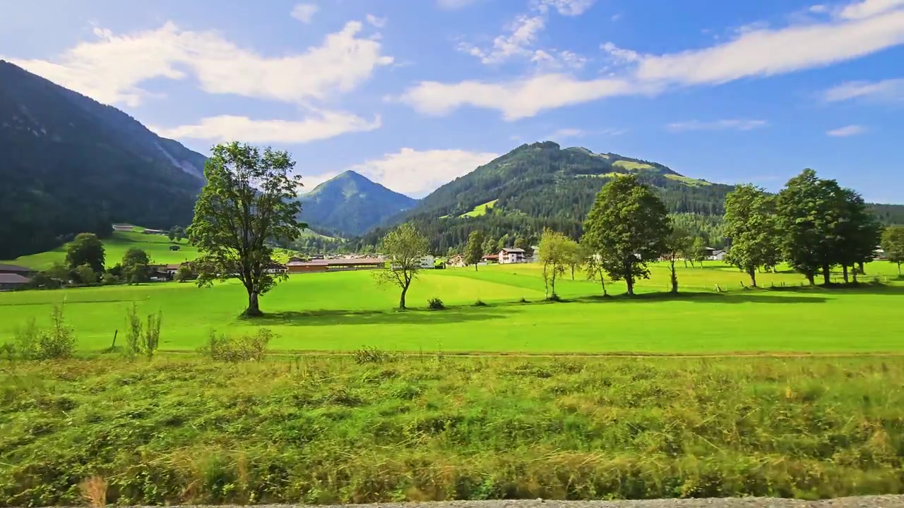 走行中の列車から見た広大なオーストリアの田園風景。豊かな緑の畑、田舎の家々、森林に覆われた尾根、そして穏やかなアルプスの谷の上に広がる青空が特徴。