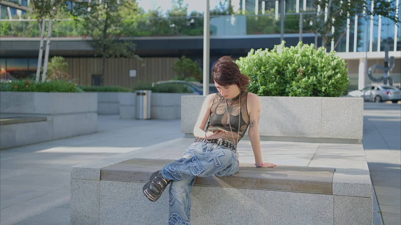 retrato de moda al aire libre de una mujer joven de estilo alternativo con tatuajes sentada en un banco enviando mensajes en el teléfono móvil 1