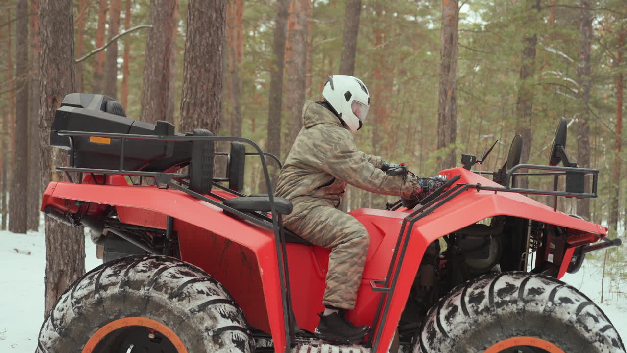 Un pilota di quad in una foresta di pini innevata accelera, giacca mimetica e casco bianco visibili, un quad rosso con pneumatici tassellati fermo tra gli alberi, primo piano delle mani sul manubrio, concentrazione tecnica e adrenalina.