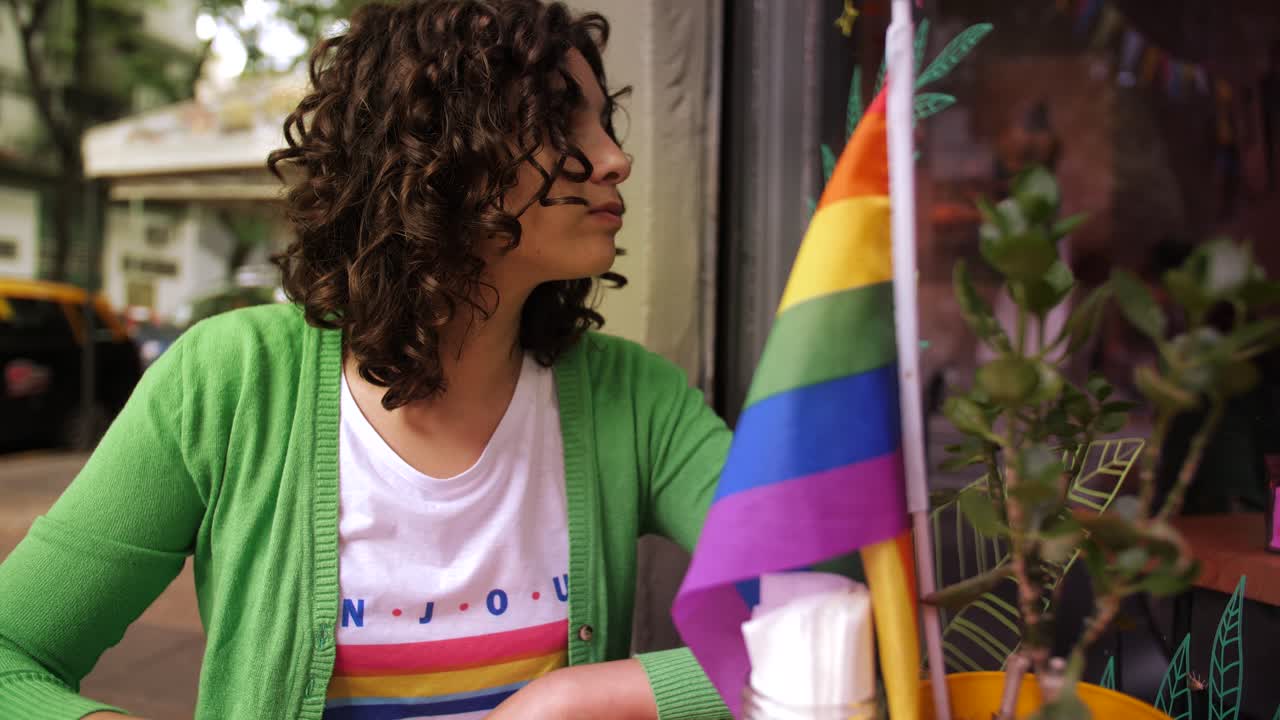 Young Woman Drinks Coffee at Outdoor Cafe with Rainbow Flag