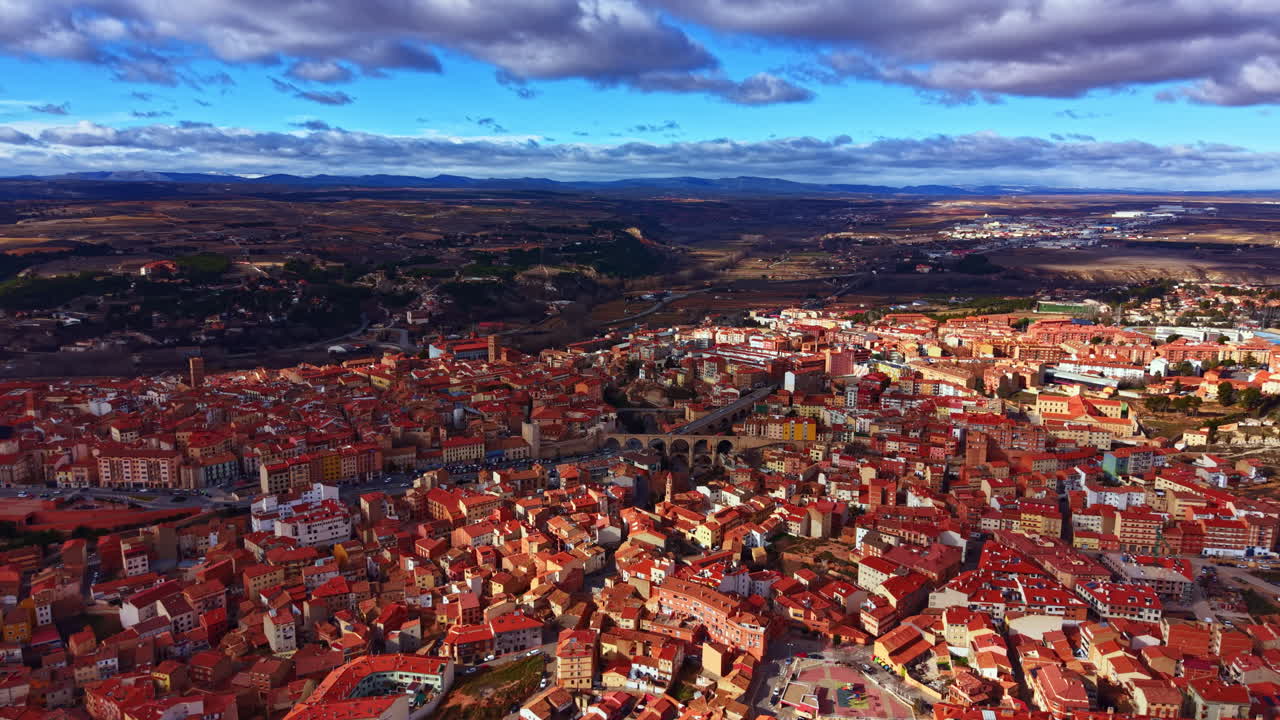 Vista aerea di una cittadina in Spagna con tetti rossi, montagne sullo sfondo e nuvole sparse in cielo, di giorno, in autunno.