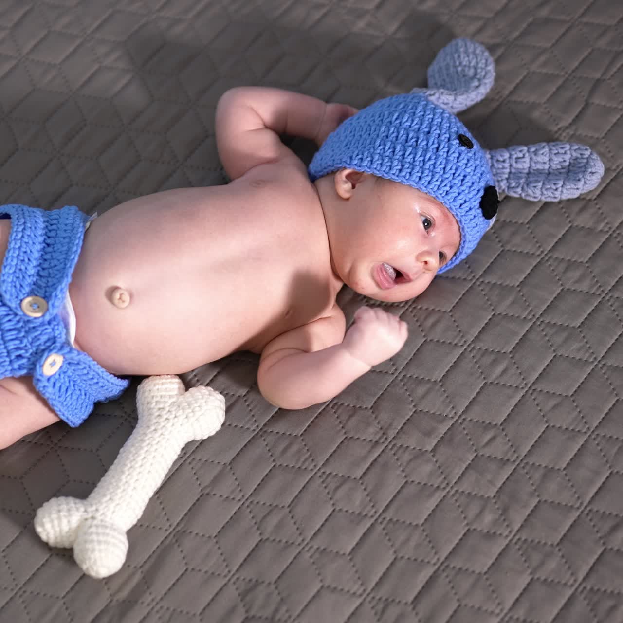 Baby boy in a knitted doggy costume lying on the bed. White toy bone beside. Cute kid at the grey backdrop