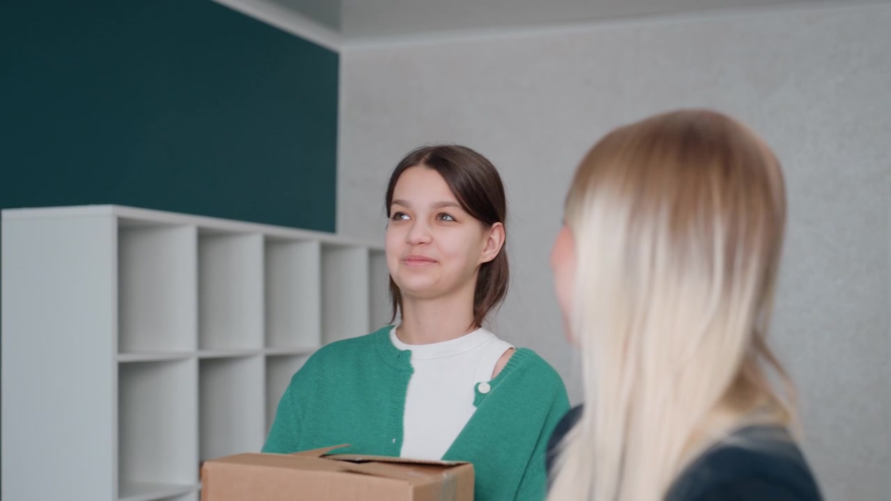 Due donne che trasportano uno scatolone all'interno di un appartamento vuoto, parete d'accento color ottanio e scaffali a cubo bianchi, la mora che tiene il cartone mentre la bionda osserva, silenzio delicato e sorrisi esitanti, piccola pianta sul bancone.