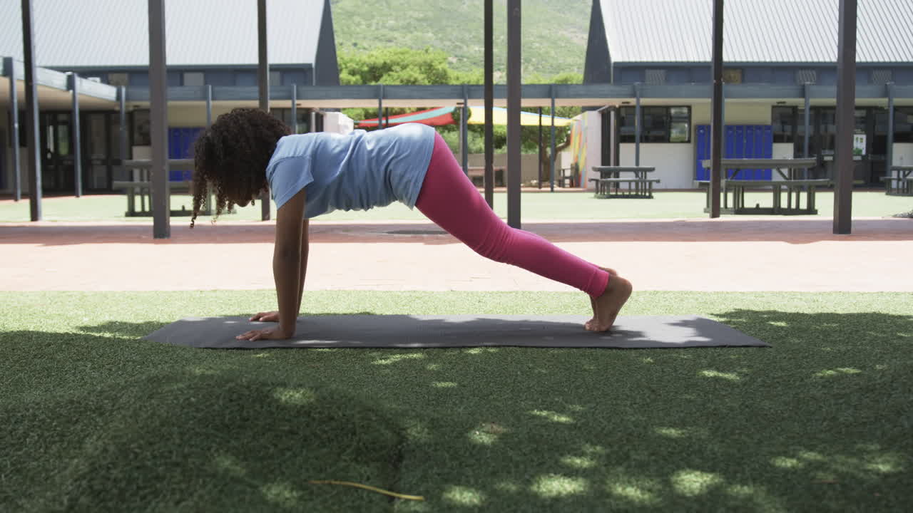 niña biracial practica yoga en una posición de tabla al aire libre en la escuela