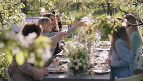 Frauen-Essen-Draußen-In-Einem-Wunderschönen-Blühenden-Garten-Zu-Mittag.-Damen-Stoßen-Gläser-Mit-Wein-An