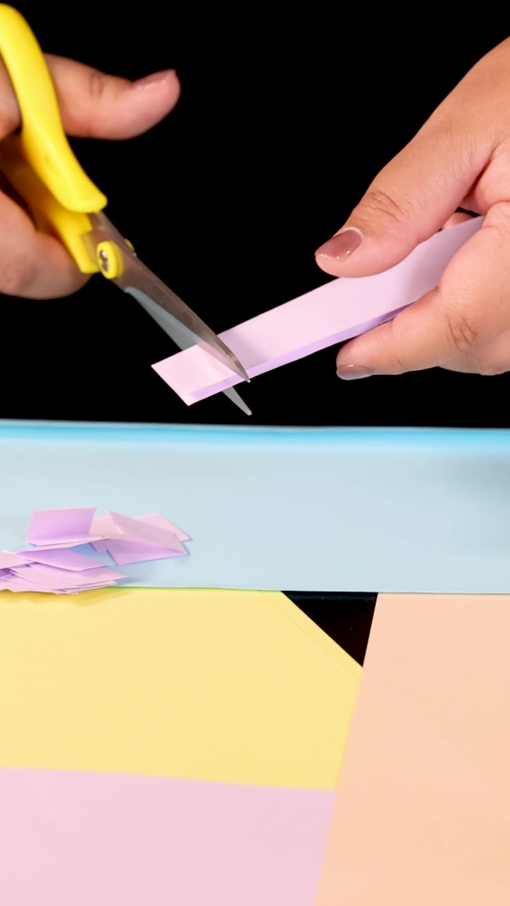 Primo piano verticale di mani che usano forbici gialle per tagliare carta rosa su sfondi pastello.