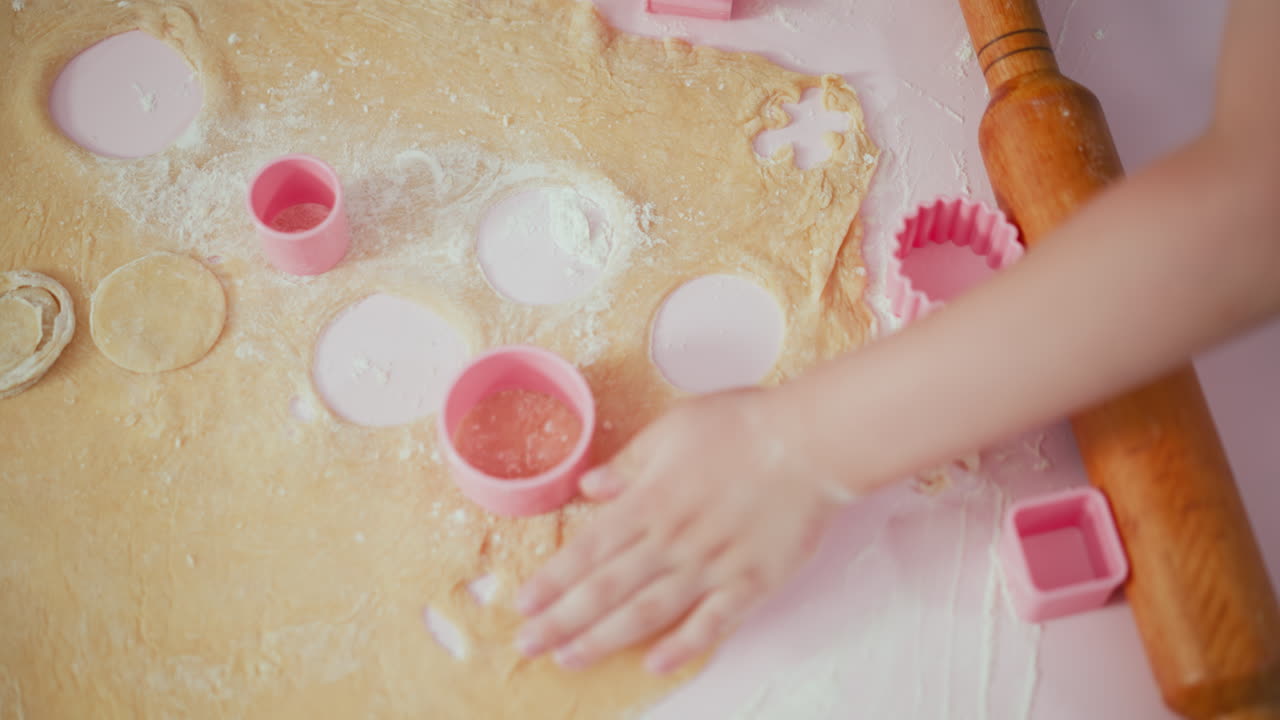 Overhead view of children pressing pink square shape cutter into flattened dough while baking, hands actively working together on floured table with wooden rolling pin and baking tools around