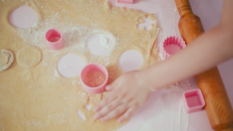 overhead view of children pressing pink square shape cutter into flattened dough while baking, hands actively working together on floured table with wooden rolling pin and baking tools around